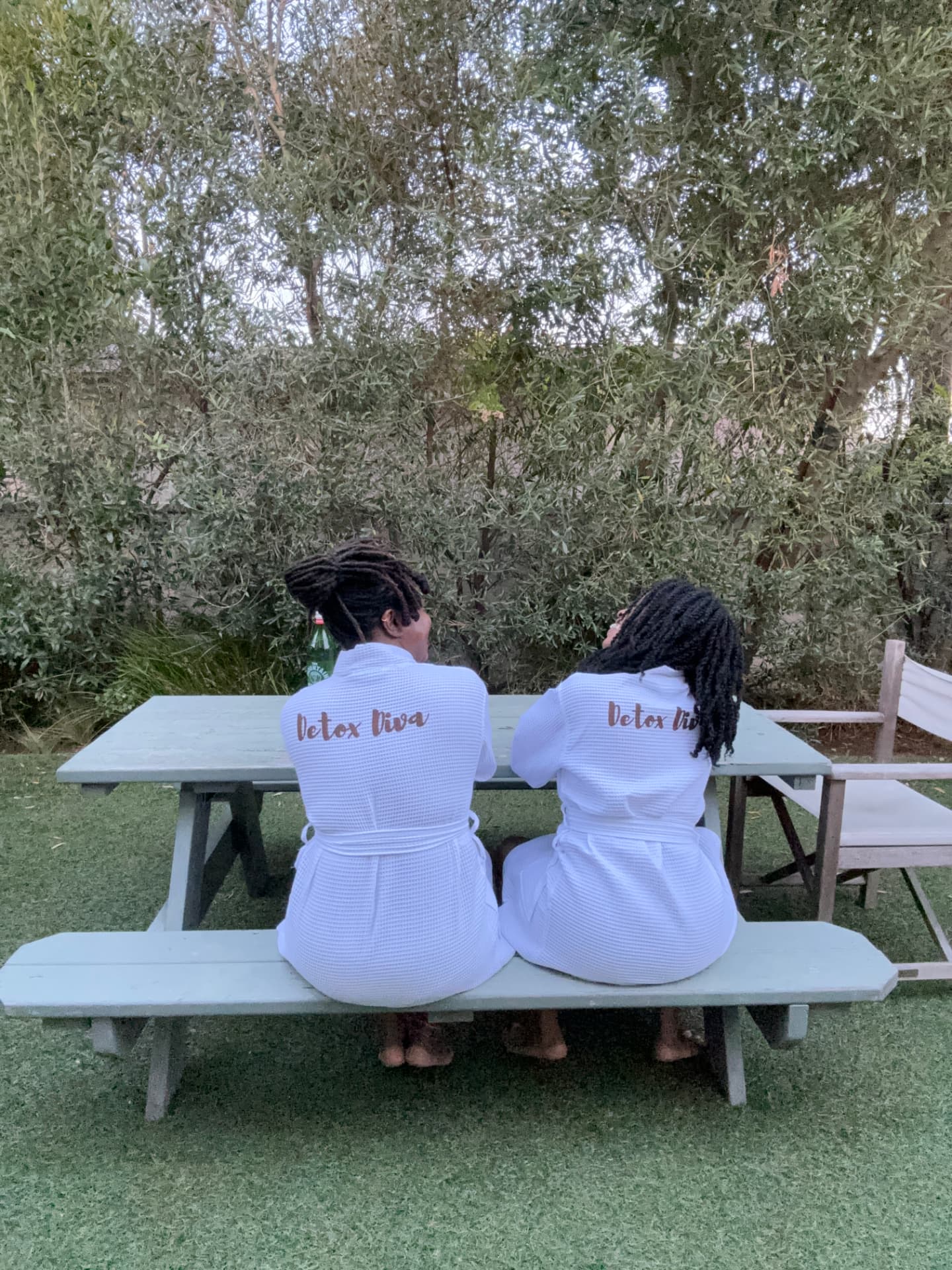 Two women in white robes sitting side by side at an outdoor table under olive trees